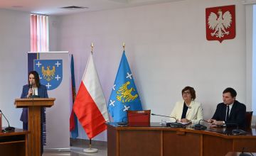 Sala konferencyjna przy mównicy stoi kobieta przy półokrągłym stole siedzą dwie osoby